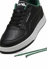 Black leather sneaker with white outsole, black laces, and green accents on the collar. Features perforations on the toe and branding on the side.