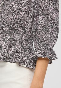 Zwart-roze gebloemde blouse met ruffles aan de mouwen, voorzien van een knoopsluiting en een subtiele merketiket aan de onderrand. Lichtgewicht stof.