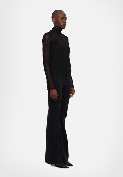 Schwarzes langärmliges Turtleneck aus durchsichtiger Stoff mit strukturiertem Blumenmuster, kombiniert mit figurbetonten, dunklen Schlaghosen und spitzen schwarzen Schuhen.