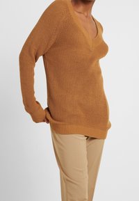 Rostfarbener Strickpullover mit V-Ausschnitt, langen Ärmeln und gerippten Säumen, kombiniert mit hellbeigen, taillierten Hosen. Texturierte Stoffdetails sind sichtbar.