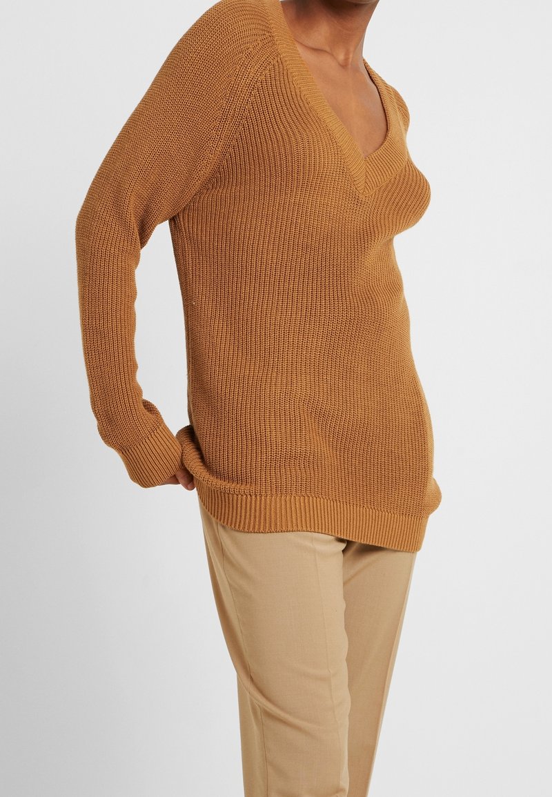 Rostfarbener Strickpullover mit V-Ausschnitt, langen Ärmeln und gerippten Säumen, kombiniert mit hellbeigen, taillierten Hosen. Texturierte Stoffdetails sind sichtbar.