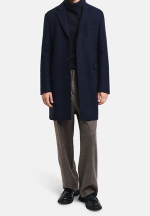 Mann in marineblauem Mantel und schwarzem Rollkragenpullover, brauner weiter Hose und schwarzen Lederschuhen, der vor einem einfarbigen Hintergrund steht.