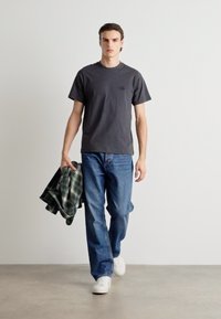 Mørk grå bomull T-skjorte med korte ermer, kombinert med blå denimbukser. Modellen holder en grønn rutete skjorte og har på seg hvite sneakers.
