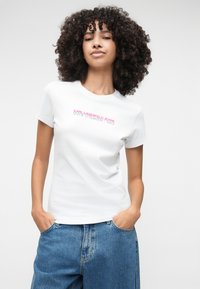 Valkoinen puuvillainen t-paita, jossa on vaaleanpunaista tekstiä "KARL LAGERFELD JEANS." Lyhyet hihat ja pyöreä pääntie, yhdistettynä sinisiin korkeavyötäröisiin farkkuihin.