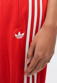 Czerwone spodnie sportowe z białymi bocznymi paskami, z małym białym logo i detalem kieszeni na zamek. Smukły krój, miękka faktura materiału.