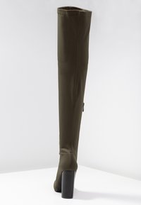 Bota verde oliva oscuro sobre la rodilla con un tejido liso y elástico, tacón cuadrado y cremallera lateral; presenta detalles de costura minimalistas.