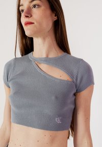 Top corto grigio a coste con maniche corte e un design asimmetrico con ritaglio vicino al collo, caratterizzato da un piccolo logo ricamato in basso.