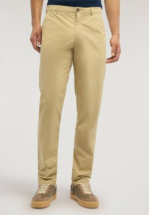 Uomo che indossa pantaloni chinos beige e scarpe da ginnastica marroni, in piedi contro uno sfondo neutro, con le mani rilassate ai lati.
