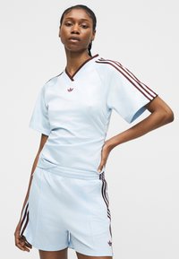 Femeie în echipament sportiv bleu deschis cu dungi bordo Adidas pe mâneci și pantaloni scurți, stă cu o mână în talie pe un fundal simplu.