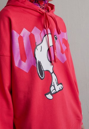Sweat à capuche rouge vif avec un grand motif blanc de Snoopy et un texte violet stylisé sur la poitrine, porté avec une écharpe colorée autour du cou.