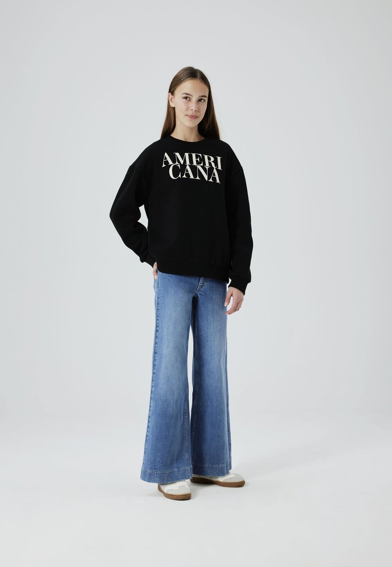 Sweatshirt noir avec le texte "AMERICANA" en blanc, associé à un jean en denim bleu évasé et des chaussures légères. Design simple, coupe décontractée.