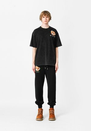 Camiseta negra de gran tamaño con un acabado texturizado y parche de oso; pantalones de chándal negros a juego con detalle en los bolsillos; botas marrones con cordones.