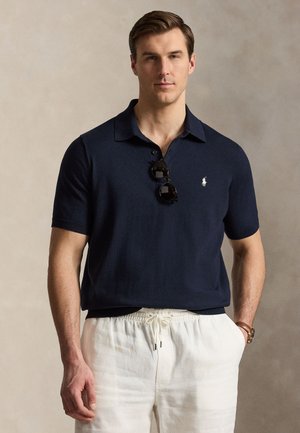 Man with short brown hair wearing a navy polo shirt with sunglasses hanging and white drawstring pants, hands in pockets.