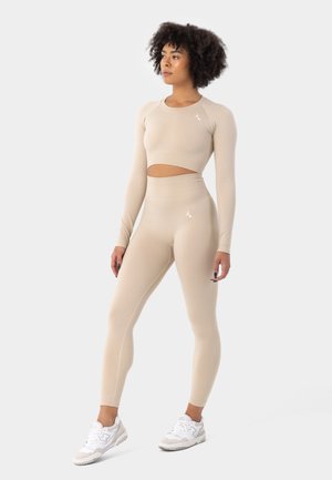 Beige treeningkomplekt, mis koosneb pika varrukaga lühikesest topsist lõhiku ja kõrge vöökohtadega retuusidest. Sile tekstuur minimaalse valge brändimisega.