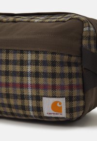 Bolsa de tela de cuadros marrones con acentos en negro, azul y rojo. Cuenta con una etiqueta blanca con un logo naranja y correas de cinta negras.