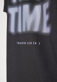 Zwarte t-shirt van katoen met grote grijze grafische tekst "TRAINING TIME" en "TRAINING CLUB 24/7" in het wit onderaan.