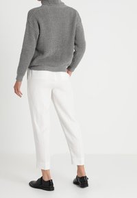 Grau gestrickter Pullover mit hohem Kragen, kombiniert mit weißen schmalen Hosen und schwarzen Schnürschuhen. Die Textur ist im Design des Pullovers ausgeprägt.