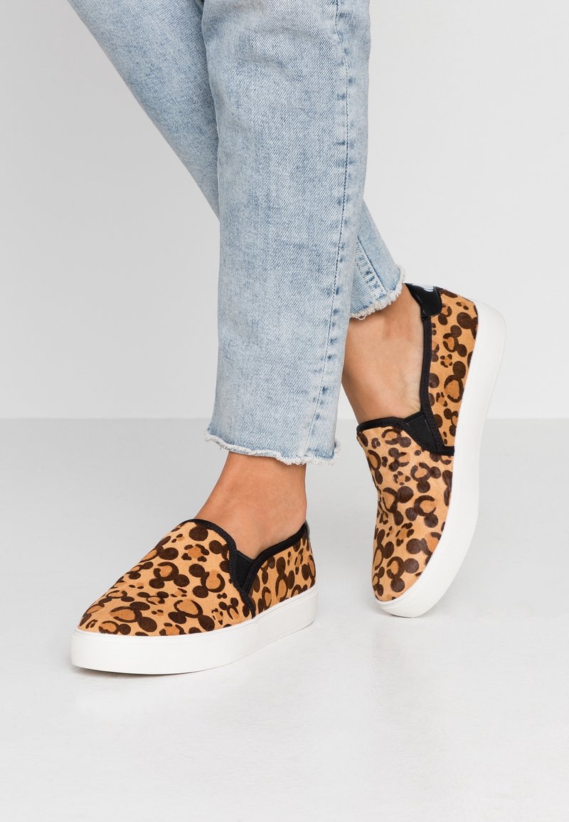 Pieds portant des baskets slip-on à imprimé léopard et bordure noire, associées à un jean court bleu clair avec des ourlets effilochés.