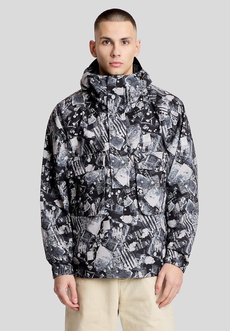 Jeune homme portant une veste à capuche à motif abstrait noir et blanc et un pantalon beige, debout devant un fond gris.