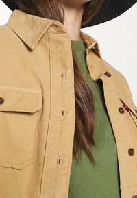 Camicia in corduroy color khaki con chiusura a bottoni, dotata di tasca sul petto e con sottogonna verde a contrasto. Tessuto texturizzato e bottoni marroni.