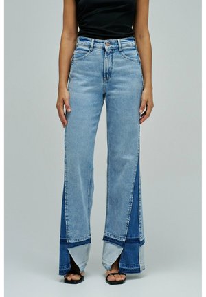 Mujer vistiendo vaqueros anchos de talle alto color azul claro con paneles de mezclilla en capas y sandalias negras abiertas en los dedos, mostrada de la cintura a los pies.