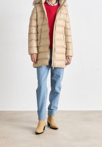 Person som bär en beige huva pufferjacka, röd tröja, blå jeans och beige ankelskor, står på ett enfärgat ljust golv mot en vit vägg.