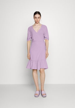 Vestido envolvente lavanda com mangas curtas e bufantes, bainha com babados e um tecido texturado. Combinado com sandálias plataforma grossas e em estilo coordenado.