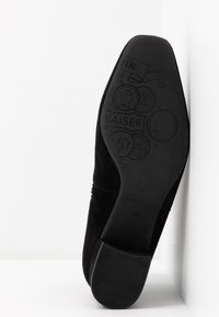 Semelle de chaussure en daim noir avec motifs de pièces embossés et texte "KAISER", reposant contre une surface blanche.