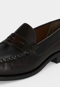 Μαύρο ανδρικό μοκασίνι από δέρμα με ραφές τύπου moc toe, λουράκι penny στο επάνω μέρος και χαμηλό ακονισμένο τακούνι σε λευκό φόντο.