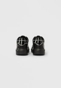 Zapatillas de charol negro con parte trasera texturizada, detalles metálicos y un logotipo. Forma redondeada con suela acolchada.