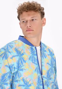 Giacca bomber leggera con sfondo blu decorato da motivi di foglie di palma gialle e rosa, rifinita con un colletto blu a contrasto e chiusura con zip.