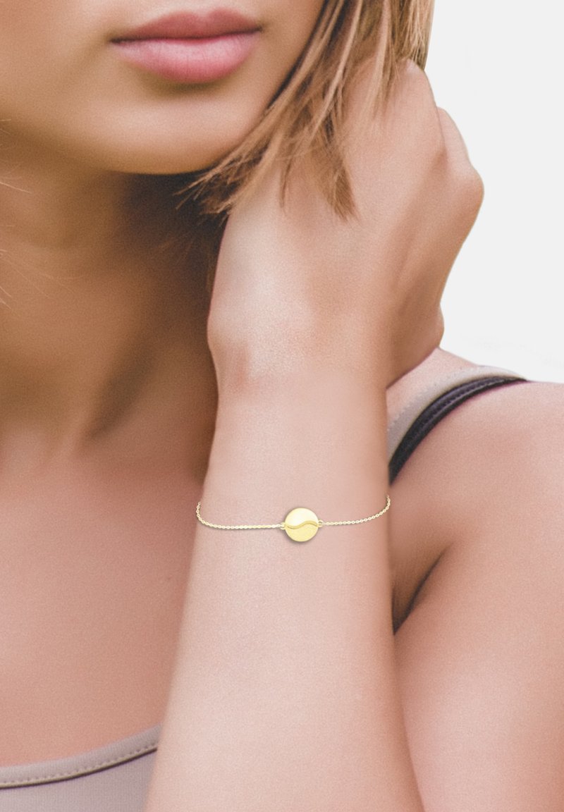 Bracciale d'oro con un ciondolo a disco rotondo su una delicata catena, indossato sul polso, su uno sfondo di tono della pelle neutro.