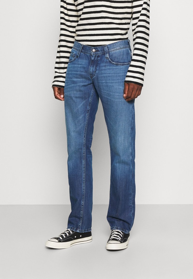 Vaqueros azul medio con corte recto, que presentan bolsillos delanteros y un sutil desgaste. Combinados con una camiseta de mangas largas a rayas en blanco y negro y zapatillas.