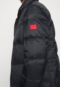 Chaqueta negra tipo puffer con diseño acolchado y un parche rojo con el logo "Hugo" en el hombro. Cuenta con cuello alto y cremallera frontal.