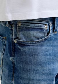 Bela majica rahlo zataknjena čez žep modrih kavbojk z vidnimi šivi in zakovicami.