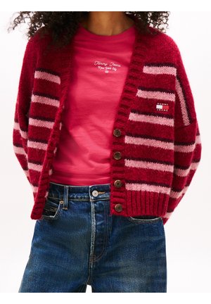 Mujer con un cárdigan de rayas rojas y rosas sobre una camiseta rosa de Tommy Jeans y vaqueros azules, mostrada desde los hombros hasta las caderas.
