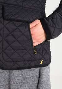 Black quilted jacket featuring a zippered pocket, textured fabric, and gold hardware accents. The inner lining is gray knit.
