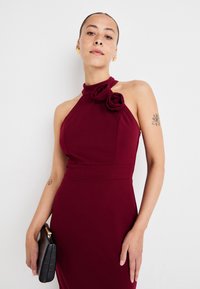 Burgundy-henkinen mekko, jossa on kukka-aukko kaula-aukossa, muotoiltu siluetti ja sileä rakenne. Musta clutch-laukku krokotiilikuviolla.