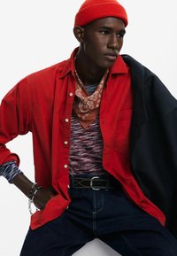 Rød skjorte med knapper over en stribet top, parret med mørk denim, et brunt bandana og en orange strikket beanie, med sølvfarvede accessories.