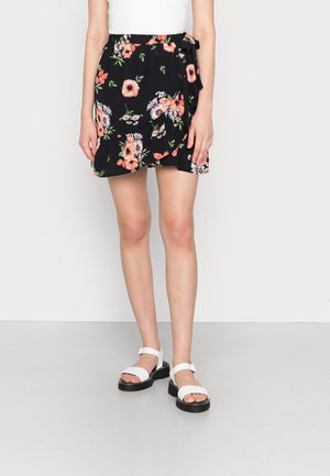 Saia envolvente preta com flores rosas e brancas, apresentando uma cintura elástica e detalhe de laço lateral. Combinada com sandálias brancas.