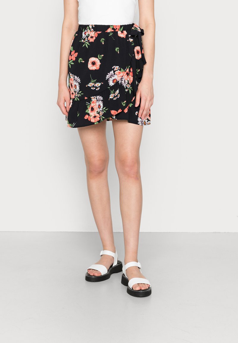 Saia envolvente preta com flores rosas e brancas, apresentando uma cintura elástica e detalhe de laço lateral. Combinada com sandálias brancas.