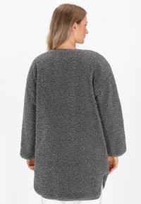 Cappotto grigio peloso con materiale testurizzato e morbido, maniche lunghe e vestibilità rilassata che arriva a metà coscia. Nessun hardware o motivo visibile.