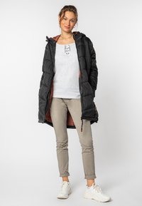 Jonge vrouw die glimlacht, gekleed in een zwarte gewatteerde jas, lichtgrijze vetershirt, beige broek en witte sneakers, staande tegen een effen achtergrond.