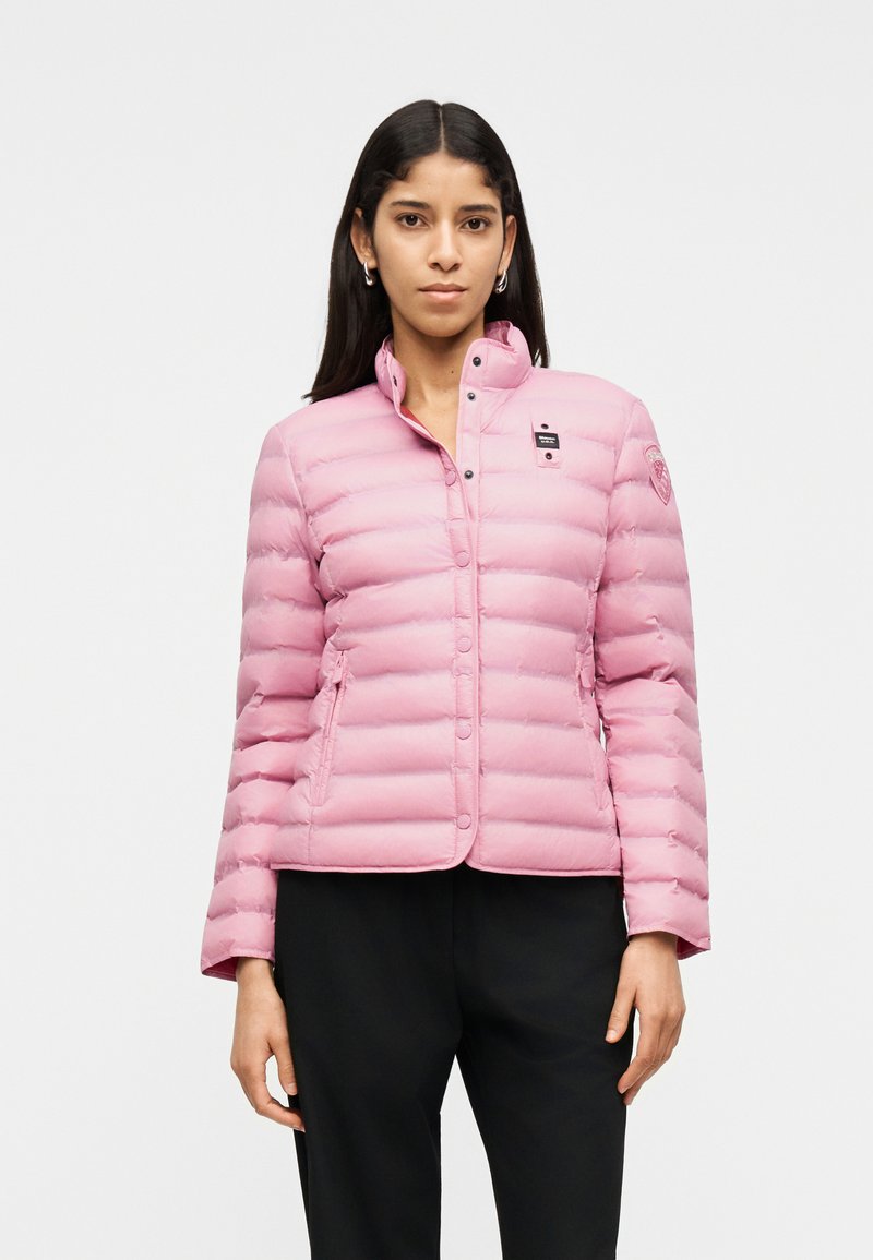 Νεαρή γυναίκα που φορά ένα ανοιχτό ροζ καπιτονέ μπουφάν με κουμπιά τύπου snap, στέκεται μπροστά σε ένα απλό λευκό φόντο.