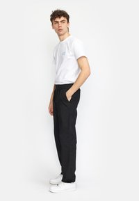 Witte katoenen t-shirt met blauwe logo, gecombineerd met zwarte broek met elastische tailleband. Model draagt witte sneakers, in een zijaanzicht.
