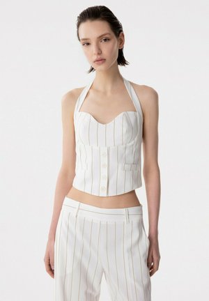 Giovane donna che indossa un top halter bianco a righe sottili con bottoni e pantaloni coordinati a vita alta, in piedi davanti a uno sfondo bianco semplice.