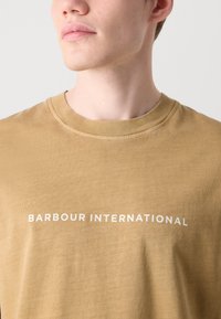 Heled pruun puuvillane t-särk ümmarguse kaelusega, millel on rinnal valged julged tähed "BARBOUR INTERNATIONAL".