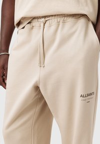 Beige sweatpants med snøre i taljen og "ALLSAINTS" logo i sort på højre lår, båret med en matchende beige top.