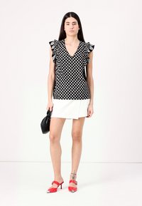 Blusa bianca e nera con pois e maniche a volants, abbinata a una mini gonna bianca e sandali con tacco rossi. Borsa nera in mano.