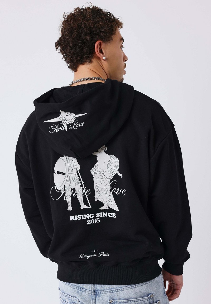 Homme portant un sweat à capuche noir avec un motif blanc représentant un guerrier ancien et une femme, texte "Antic Love", "Rising Since 2015" et "Design à Paris".